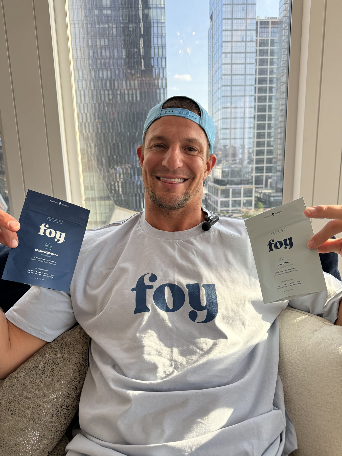 A smiling man wearing a light blue Foy cap and a white t-shirt with a blue Foy logo sits on a couch holding two Foy product packets, one blue and one white, in front of a window with a city view.